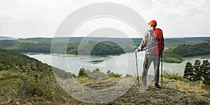 Hiker man with backpack and trekking poles standing on top of cliff and looking at the mountains.