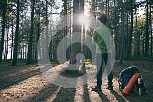 Hiker man in felt hat with backpack and Ñamping gear standing in pine woods at sunset. Adventure, trekking, local travel. Copy