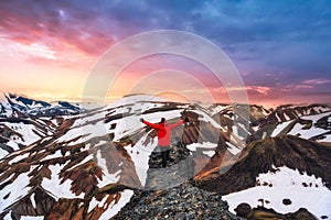Hiker man standing with arms raised on the cliff with volcanic mountain and snow covered view in summer at Landmannalaugar