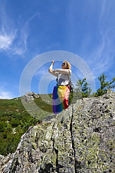 A hiker perched on a rock at the top of a mountain, with a rainbow flag hanging from her waist and making a sleeve cut