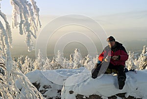 A hiker pours himself tea on top of a cliff in winter