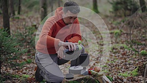 Hiker prepares food and pours water from bottle into pot. Traveler in forest cooks food with small cooking stove. Man