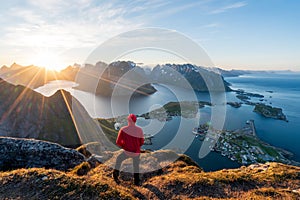 Hiker in red jacket are standing on top of the famous Reinebringen overlooking Reine and Hamnoy in the Lofoten islands during the photo