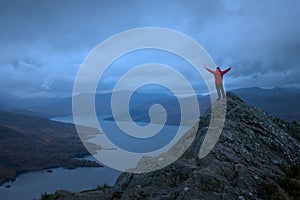 A hiker in a red jacket on top of a mountain looks out over the magnificent scenery of a lake surrounded by mountains