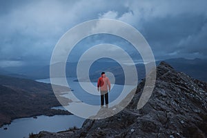 A hiker in a red jacket on top of a mountain looks out over the magnificent scenery of a lake surrounded by mountains