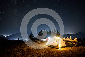 Hiker sitting near illuminated camp tent under night starry sky.