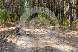 Hiker sitting on a sandy roadside and looking in backpack in  mixed forest