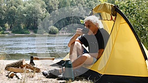 Hiker sitting in a tent. Camper man drinking coffee or tea. River and forest in the background. Relaxation, travel