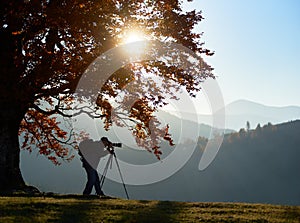 Hiker tourist man with camera on grassy valley on background of mountain landscape under big tree.