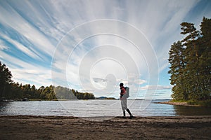 Hiker wearing a jacket and carrying a backpack walks along the beach and watches Lake Jatkonjarvi at sunset in Koli National Park