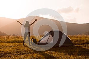 Hiker woman silhouette with backpack and hands up standing near tent and looking to mountains sunset