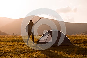 Hiker woman silhouette with backpack and hands up standing near tent and looking to mountains sunset