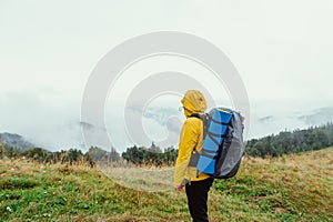 Hiker in a yellow jacket stands in the mountains on a rainy foggy day during a hike, looks at the beautiful views of nature and
