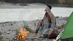 hiker young tourist traveler man enjoying nature, sitting near bonfire at mountain camping camp tent