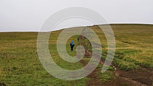 Hikers with backpacks climb a hill covered with grass. People walk along the dirt road. In the background is a gray cloudy sky. Th