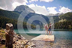 Hikers on wooden jetty by lake jumping in joy. Summer trip in nature. Lifestyle, love, togetherness, nature concept