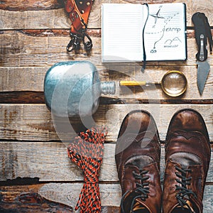 Hiking accessories on wooden background: old hiking leather boots, vintage film camera, travel notebook, knife. Lifestyle concept