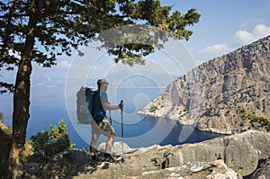 Hiking on Lycian way. Man with backpack enjoys view of Butterfly Valley blue lagoon from rocky cliff in shadow