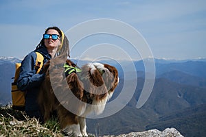 Hiking with pet. Happy young Caucasian woman with dreadlocks sitting on hill with dog. Snowy peaks of Caucasus Reserve