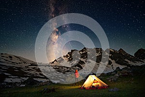 Hiking tourist standing near illuminated tent under milky way stars