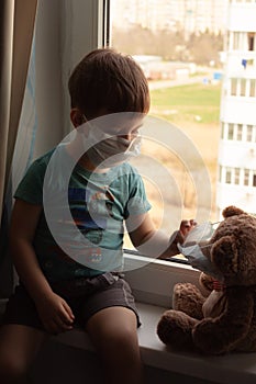 Ð¡hild and his teddy bear both in protective medical masks sits on windowsill