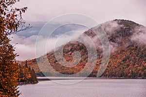 Hilly mountains in the fog covered with autumn bright forest on the shore of the lake. autumn gloomy day.