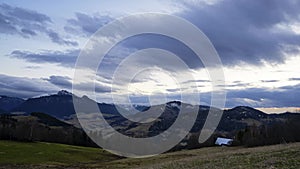 A hilly rural landscape, clouds moving at dusk, grassy meadows with trees, mountains,