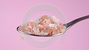 Himalayan coarse rock salt in a spoon on a colored pink background. Falling orange rough crystals.