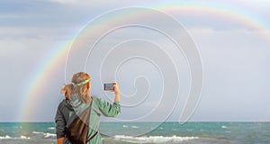A hippie blogger girl takes pictures of a rainbow on a mobile phone. Beauty over the sea. The concept of travel