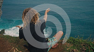 Hippie man with long curly hair sitting in lotus position on edge of ocean cliff moving hands as if controlling power of