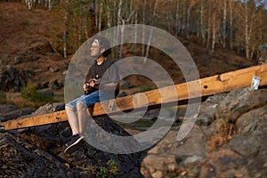 a hippie man sits on a bridge near the rocks and plays the ukulele