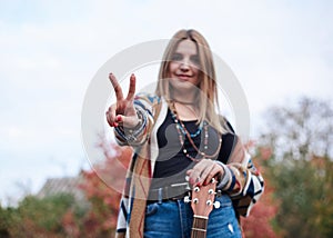 Hippie musician with acoustic guitar, showing piece sign with two fingers. Three-quarter portrait of young woman, wearing colorful