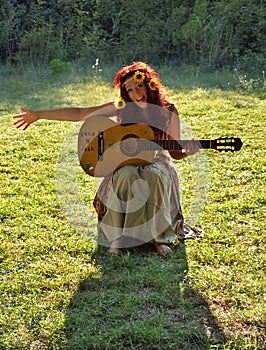 Un bel sorridente bruna con un fiore nei capelli, seduta sull'erba verde a suonare la chitarra e a cantare una canzone.