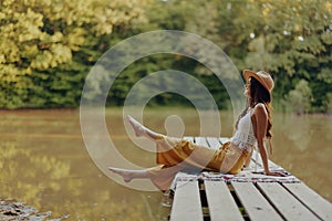 A hippie woman sits on a bridge by the river and enjoys the beautiful scenery around her cheerfully lifting her legs