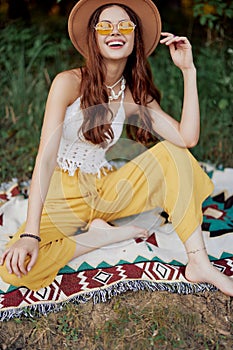 Hippie woman smiling in eco clothing yellow pants, white knit top, hat and yellow glasses sitting on plaid in park