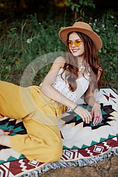 Hippie woman smiling in eco clothing yellow pants, white knit top, hat and yellow glasses sitting on plaid in park