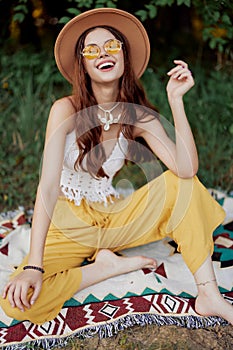 Hippie woman smiling in eco clothing yellow pants, white knit top, hat and yellow glasses sitting on plaid in park