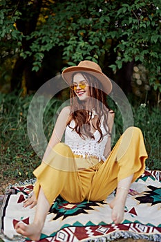 Hippie woman smiling in eco clothing yellow pants, white knit top, hat and yellow glasses sitting on plaid in park