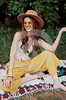 Hippie woman smiling in eco clothing yellow pants, white knit top, hat and yellow glasses sitting on plaid in park