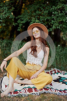 Hippie woman smiling in eco clothing yellow pants, white knit top, hat and yellow glasses sitting on plaid in park
