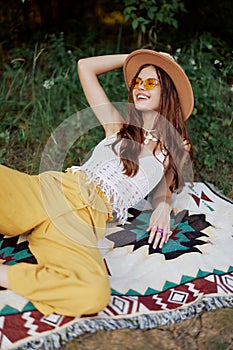 Hippie woman smiling in eco clothing yellow pants, white knit top, hat and yellow glasses sitting on plaid in park