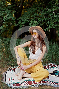 Hippie woman smiling in eco clothing yellow pants, white knit top, hat and yellow glasses sitting on plaid in park