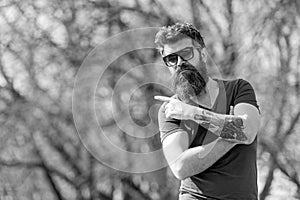 Hipster with beard looks stylish on sunny day. Man with beard and mustache on strict face pointing, nature background