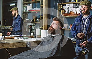 Hipster bearded client got hairstyle. Barber with hairdryer blows off hair out of cape. Barber with hairdryer works on