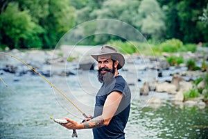 Hipster bearded man catching trout fish. Young man fishing. Fisherman with rod, spinning reel on river bank. Man