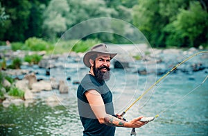 Hipster bearded man catching trout fish. Young man fishing. Fisherman with rod, spinning reel on river bank. Man