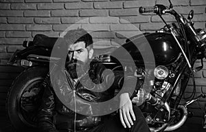 Hipster, brutal biker on pensive face in leather jacket sits, leans on motorcycle. Bikers lifestyle concept. Man with