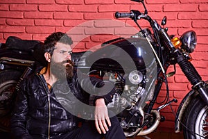 Hipster, brutal biker on pensive face in leather jacket sits, leans on motorcycle. Man with beard, biker in leather