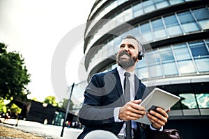 Hipster businessman with headphones and tablet sitting on a bench on the street in London.