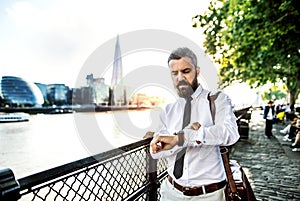 Hipster businessman with laptop bag walking by the river in London, checking the time.
